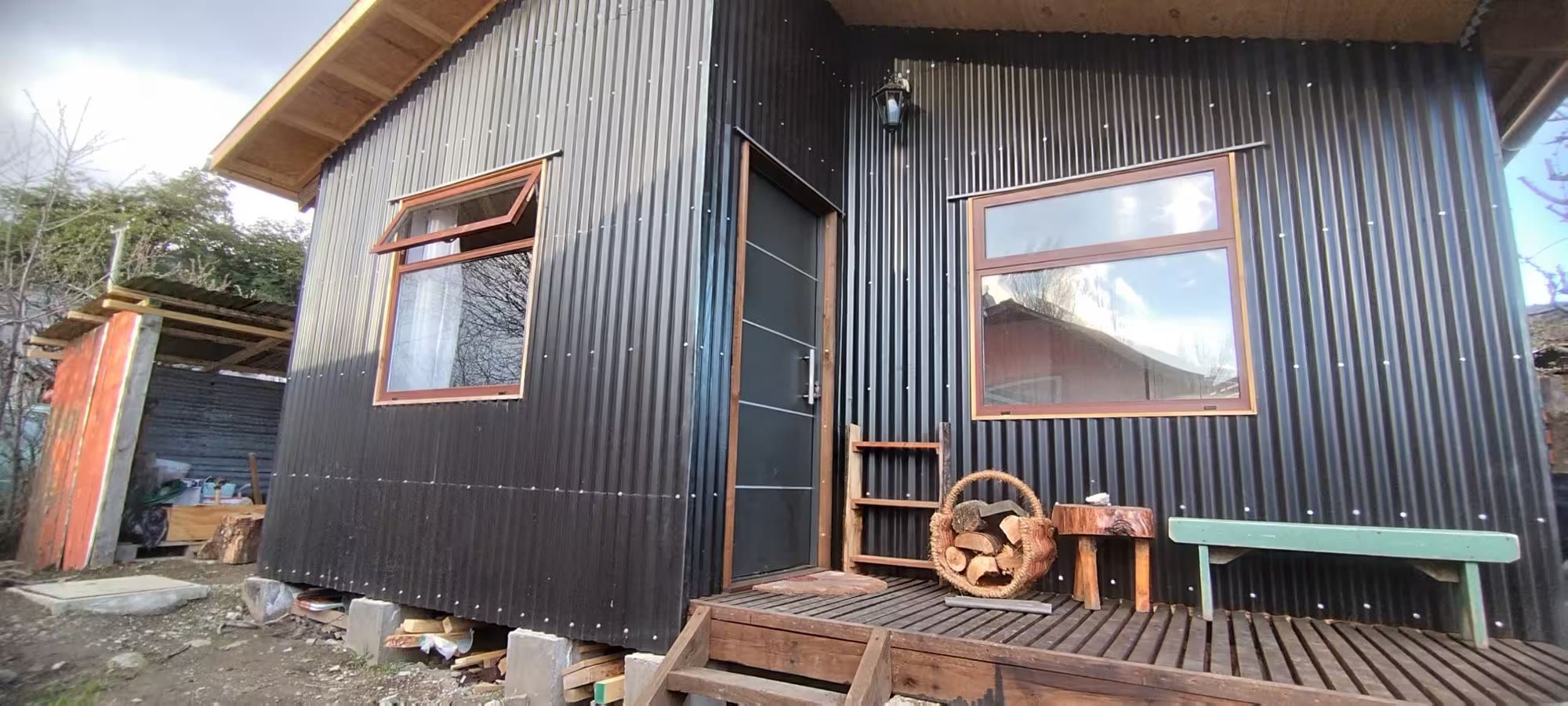 La fachada de zinc negro de La Casa del Fondo, con deck de madera, leña apilada y una banca verde